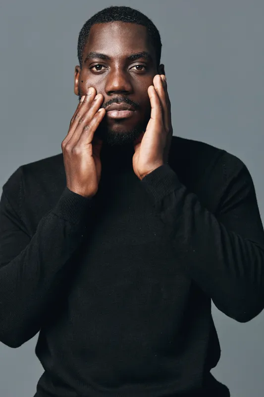 African man with dark skin in black sweater touching face with hands in creative conceptual visual style studio portrait. Serious expression and direct gaze on gray background.