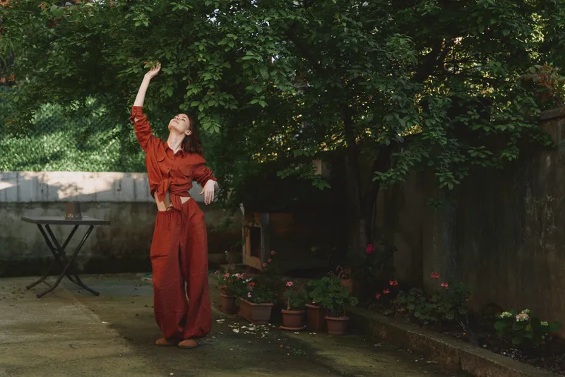 Vibrant woman outdoors in a red outfit, mid dance with raised arm, sunlit garden background, candid lifestyle moment, relaxed posture, warm tones, lush greenery
