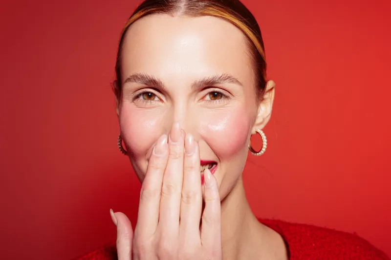 Smiling woman with clear skin covers mouth with hand on red background. Close-up portrait with hoop earrings and natural makeup. Concept of happiness, beauty, and positive emotion.
