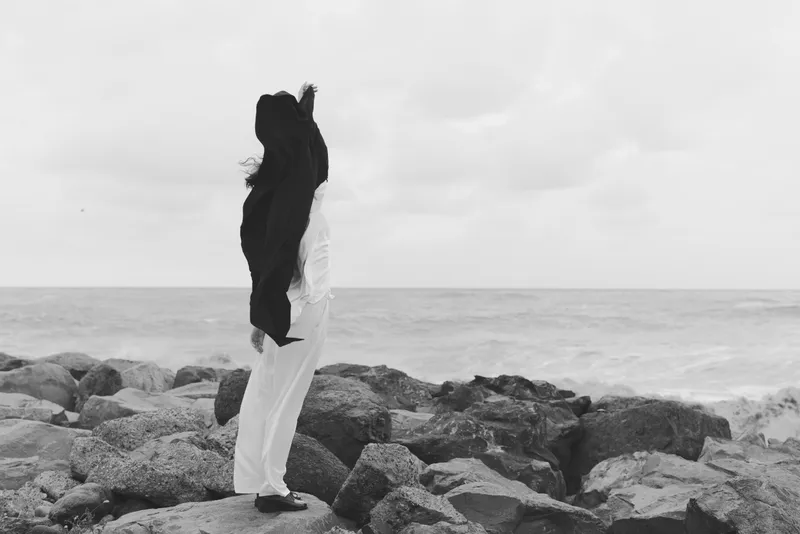 Couple embrace on a rocky coast with ocean in the background, wedding attire flowing, black and white scene capturing romance, memory and quiet togetherness