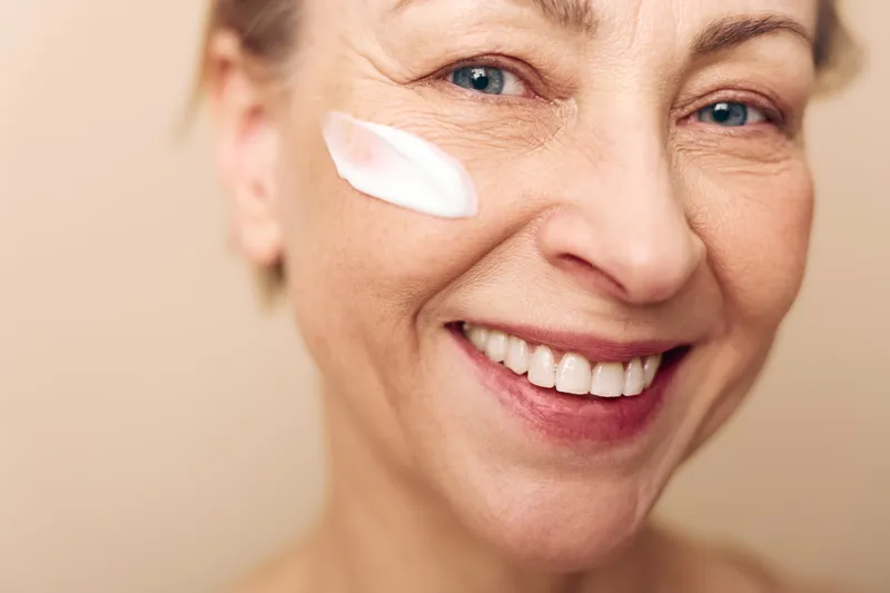 Close-up of happy mature woman with blue eyes applying white face cream on cheek. Concept of skincare, antiaging, natural beauty, wellness, and self-care in soft studio lighting.