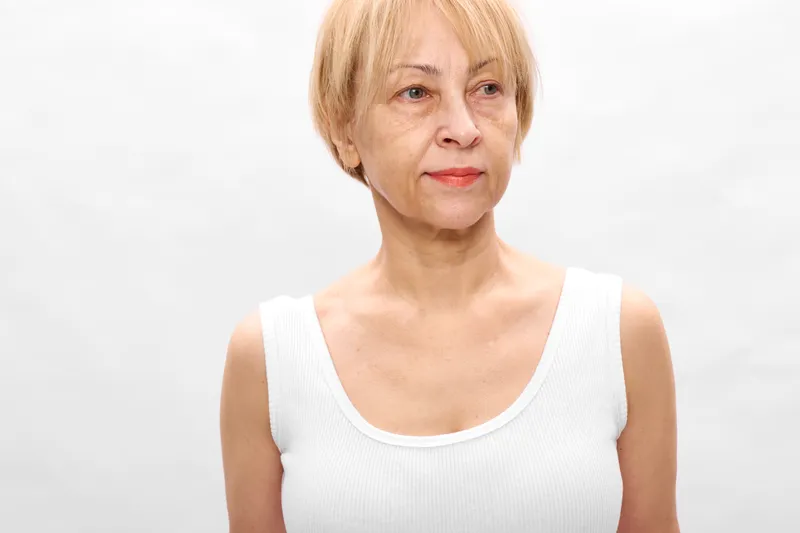 Older woman portrait closeup in studio, senior female, short blonde hair, white sleeveless top, neutral expression, isolated on clean white background.