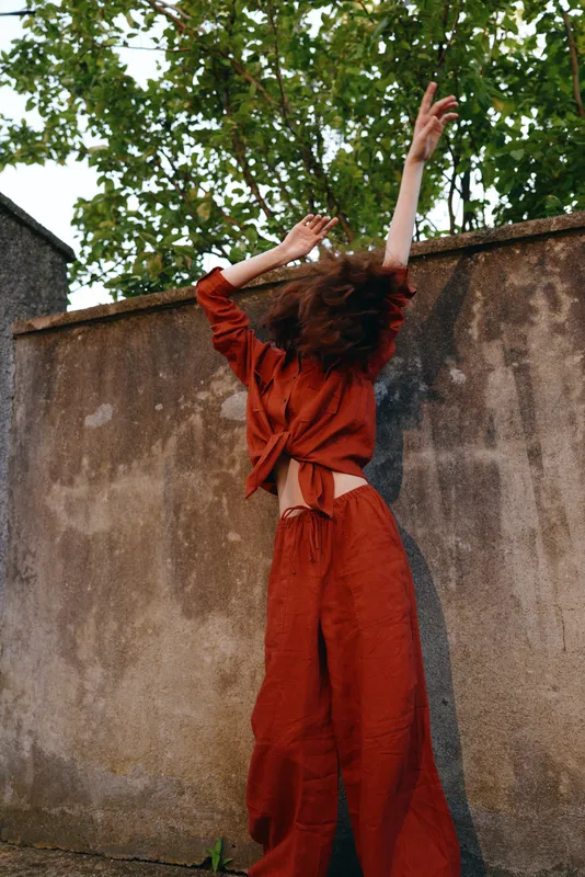 A young woman in a red jumpsuit raises her arms against a weathered wall, creating a dynamic, energetic moment that emphasizes movement and contrast in a gritty outdoor setting
