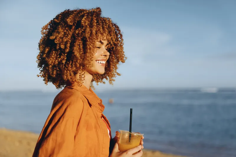 Young woman with curly hair wearing an orange shirt smiles on a sunlit beach, holding a cold drink with a straw as waves roll in and a clear blue sky frames the peaceful seaside moment, radiating