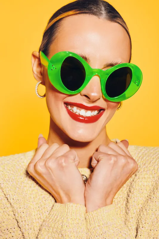 Happy woman with bright red lipstick wears large green sunglasses and yellow sweater, smiling with clenched fists on yellow background. Concept of joy, fashion, and summer style.