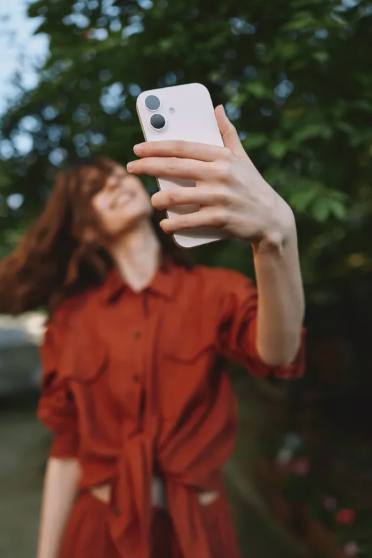 A woman takes a carefree selfie outdoors in a red dress, capturing a casual lifestyle moment in a sunlit garden. A playful vibe of modern youth and self expression