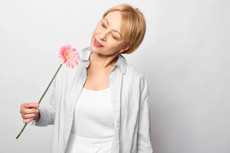 Woman studio closeup portrait holding a pink dandelion flower, smiling with eyes closed, soft gray background, spring beauty mood, casual robe shirt