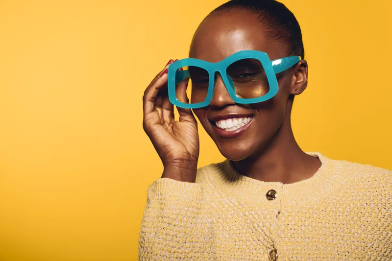 Smiling African woman with dark skin wearing oversized blue glasses and yellow sweater poses against yellow background. Concept for fashion, joy, and vibrant lifestyle with stylish eyewear.