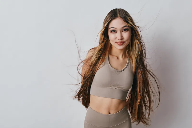 Young woman standing with long hair flowing, wearing neutral colored sportswear and smiling confidently against white background for fitness and lifestyle.