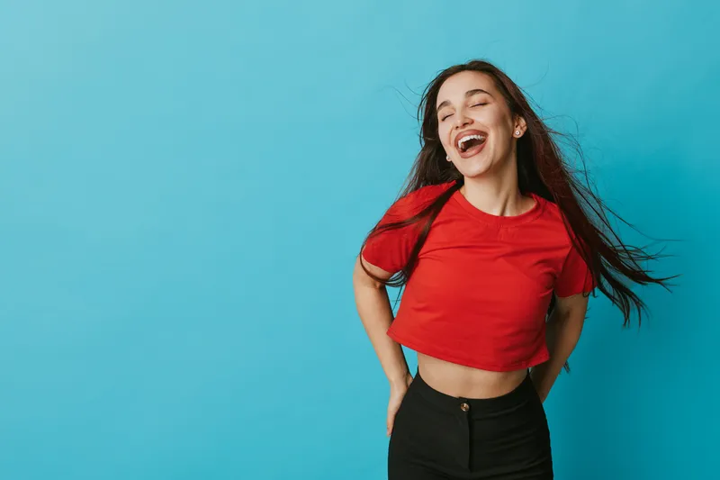 Young woman laughing with closed eyes wearing red crop top and black pants standing against blue background. Casual style, happy expression and carefree mood with long hair flowing.