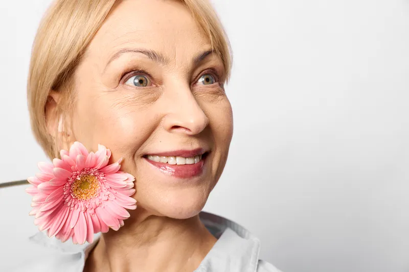 Woman with blonde hair holding a pink flower near her cheek, smile, close up portrait on a bright white background, beauty and skincare care, soft studio lighting
