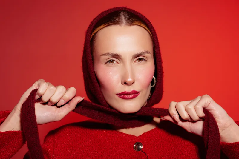 Portrait of confident woman in red sweater and hood posing against red background. She holds hood strings with both hands, looking directly at camera with calm expression.