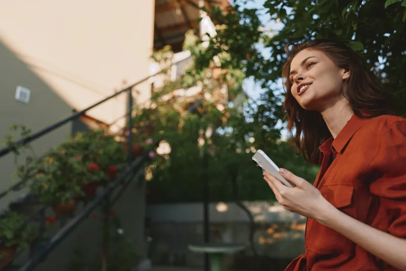 Young woman with brunette hair in an orange shirt holds a smartphone outdoors, smiling in a relaxed garden setting, with soft natural light highlighting a friendly, approachable mood