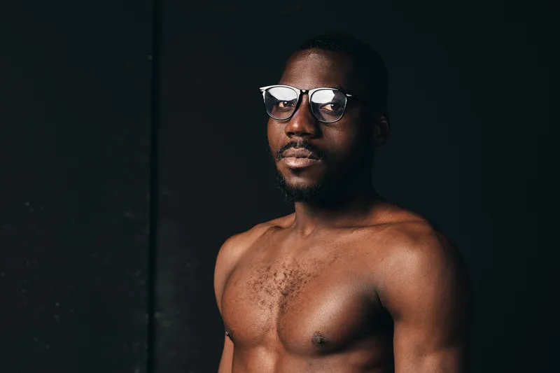 Creative conceptual portrait of a shirtless African man wearing glasses with confident expression in dark studio. Visual style emphasizes skin texture and thoughtful mood in artistic lighting.