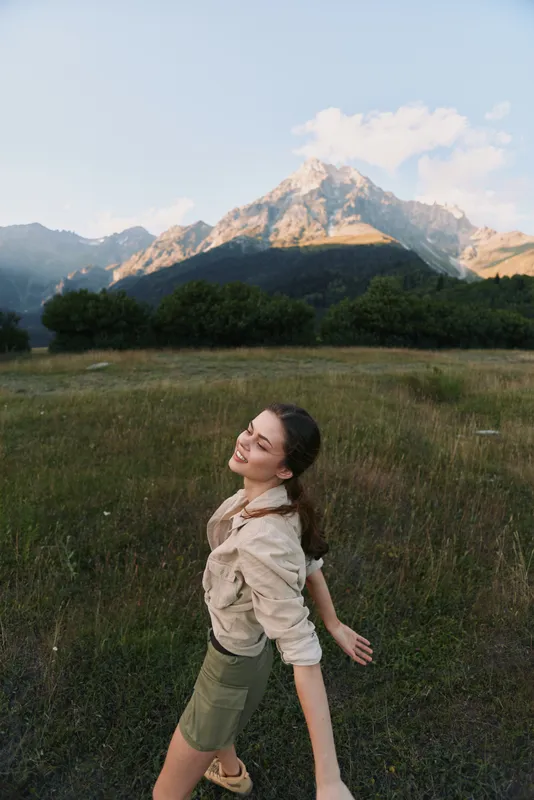 Young woman in a beige shirt and green shorts stands in a grassy field with snow capped mountains behind, enjoying sunlit freshness