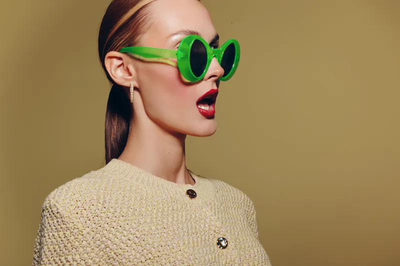 Stylish woman in green sunglasses and beige textured jacket looks surprised with open mouth on plain background. Concept of fashion, retro style, and expressive emotions in studio portrait.