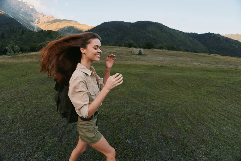 Cheerful woman outdoors, running across a grassy field with a backpack, sunlit landscape and distant mountains, carefree mood, natural colors.
