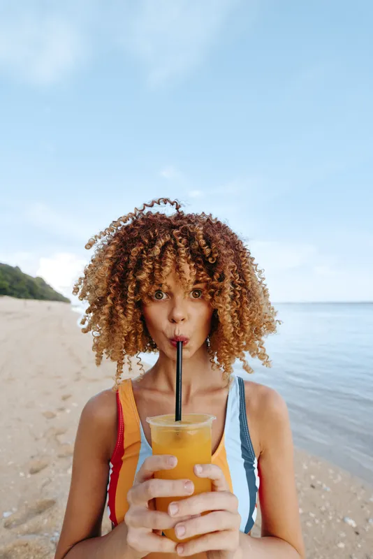 Woman with curly hair on a beach holding a juice drink with straw, summer vacation vibe, smiling closeup outdoors by ocean sand sunlight, refreshment and hydration wellness, selfcare