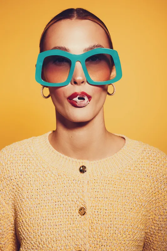 Young woman with oversized turquoise sunglasses and hoop earrings wears yellow knitted sweater, blowing bubble gum. Bright yellow background creates playful and trendy fashion concept.