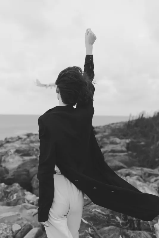empowerment, triumph, determination, gesture, a figure stands on a windy coastline with cape flowing, monochrome scene showing resilience and freedom against sea and horizon