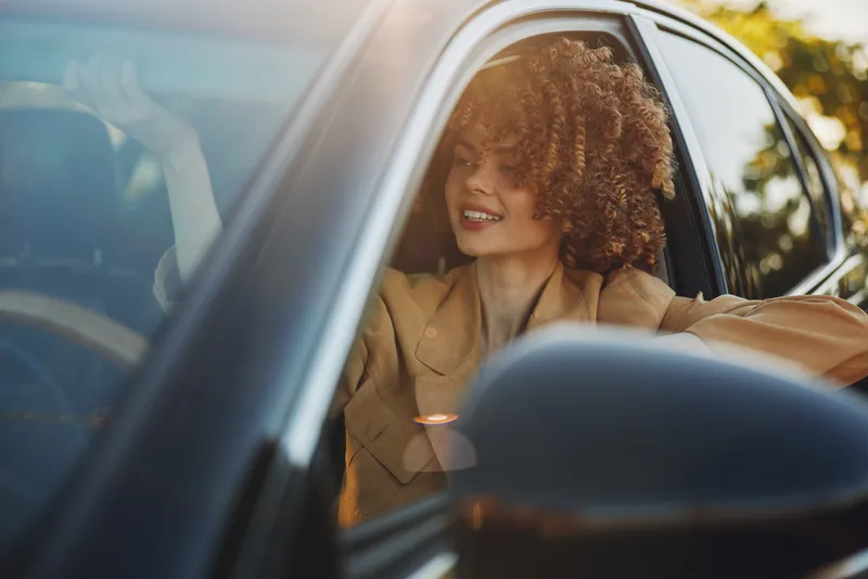 Smiling woman with curly hair sits in a car, sunlight glints on the window, warm retro tones create a relaxed lifestyle mood and sense of casual travel
