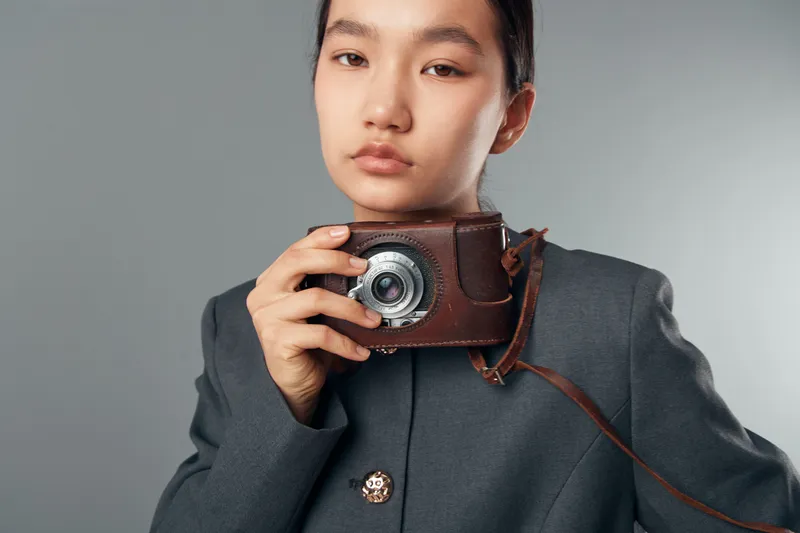 Young woman holding vintage leather camera case with camera inside, wearing dark gray coat with metal buttons, standing against plain background in studio portrait.