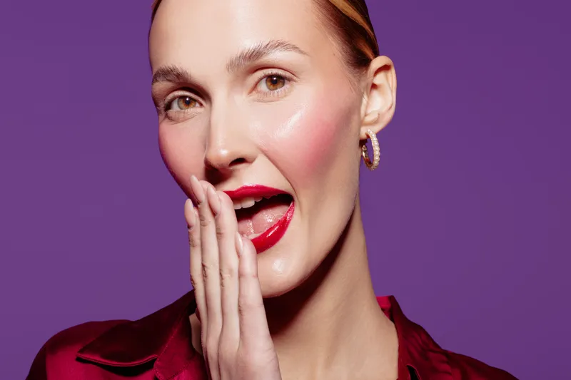 Close-up of a young woman with red lipstick and shiny skin whispering with hand near mouth on purple background. Concept of beauty, makeup, communication, and expression.