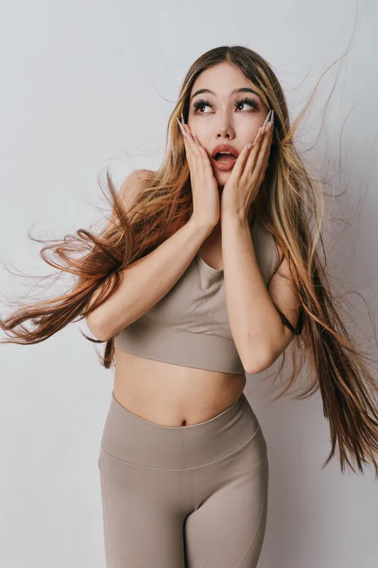 Young woman with long flowing hair poses with hands on cheeks and surprised expression in casual outfit against plain white background in studio environment.