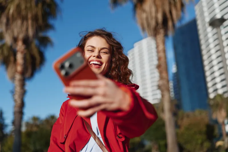 A woman in a red jacket takes a bright selfie in a sunny outdoor urban setting with palm trees and tall buildings, capturing casual city vibes and cheerful energy.