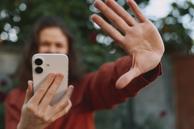 Young woman outdoors records a candid selfie with a smartphone, reaching toward the lens with a playful gesture and a relaxed smile in natural sunlight, in a sunlit garden with greenery and a soft