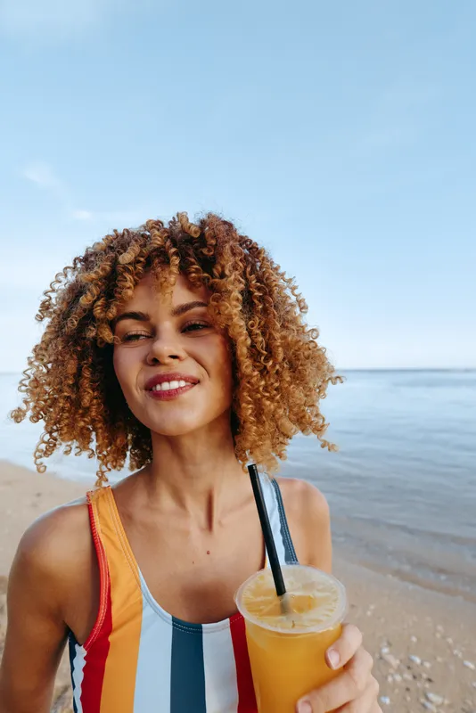 A young woman with curly hair wearing a colorful striped swimsuit enjoys a cold drink on a sunny beach with calm sea and clear sky in the background.
