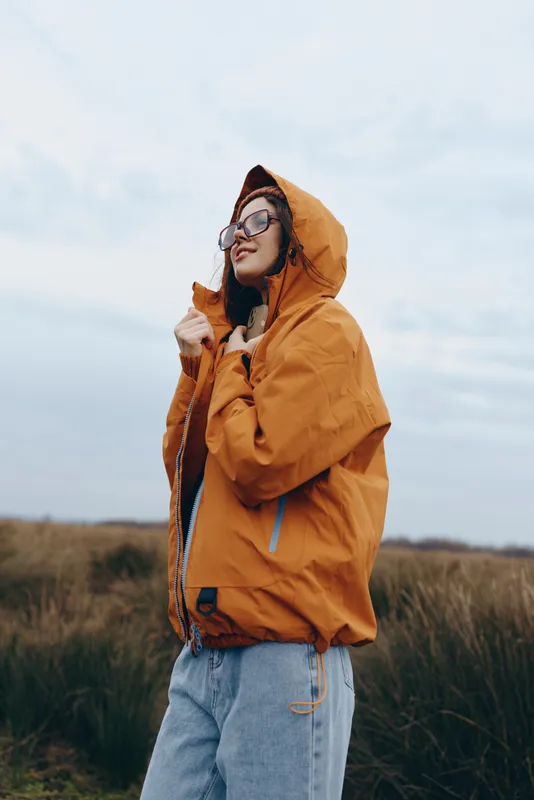 Woman lifestyle portrait outdoors in autumn field, wearing hooded jacket and glasses, standing and adjusting hood, calm mood, soft natural light, open sky background, wide meadow
