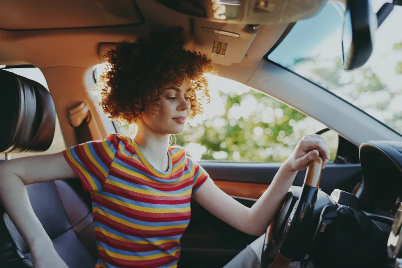 Woman driver in a striped shirt sits behind the wheel, relaxed and focused, capturing casual lifestyle with warm retro film color inside a sunlit car interior.