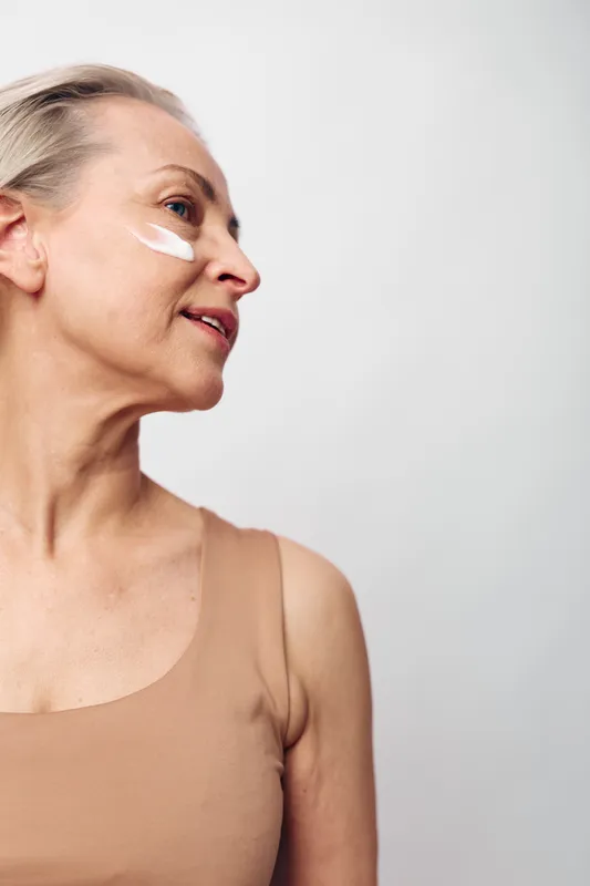 Mature woman with short blonde hair applies white face cream on cheek, wearing beige top. Skincare and self-care concept focusing on natural beauty and healthy aging in soft lighting.