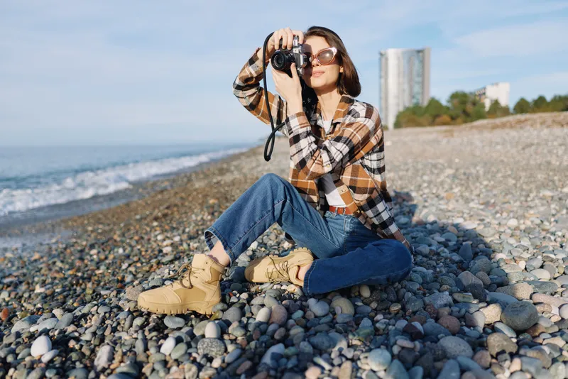 Photographer on a rocky beach with camera raised, wearing a plaid shirt and boots, capturing coastal scenery under a bright sky with distant buildings.