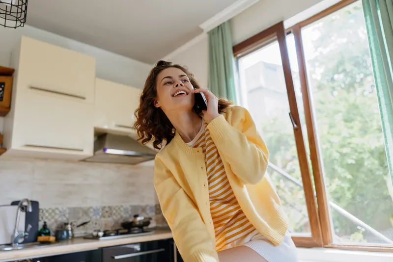 Young woman in a bright kitchen wears a yellow cardigan and striped tee, talking on a phone with a cheerful smile and relaxed posture.