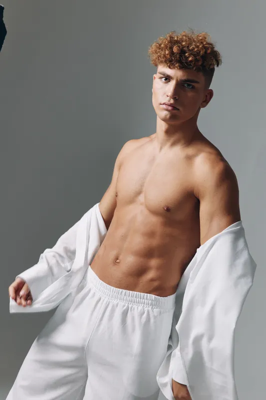 Young man with curly hair posing with shirt off and sweatpants on gray background studio light. Fitness model, fashion, bare torso with serious expression and casual relaxed stance for lifestyle.