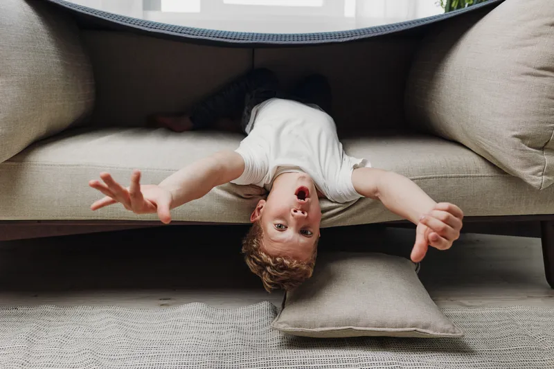 Playful boy with curly hair lying upside down under beige sofa reaching out with hands, showing joyful expression in cozy living room with natural light and soft cushions.