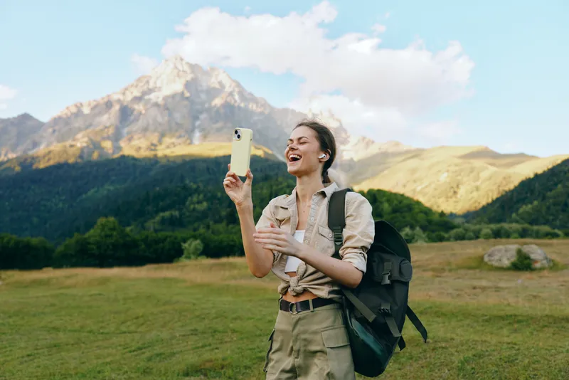 Happy woman hiking outdoors in mountains, holding smartphone, wearing backpack and casual clothes, enjoying nature, sunny day, lifestyle concept, natural landscape background