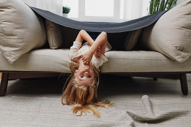 Playful girl with long hair hangs upside down from sofa covered with blanket in bright living room. Concept for childhood fun and indoor activity.