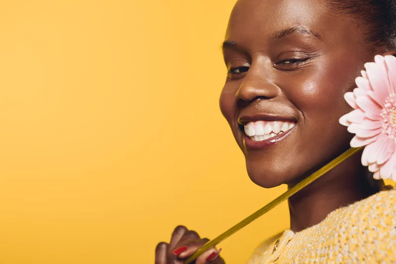 Smiling African American woman with dark skin holds pink flower on yellow background. Close-up portrait of happy female model in yellow sweater. Concept of beauty, joy, and natural elegance.