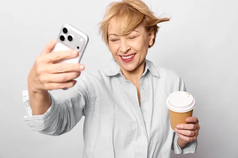 Elderly woman making selfie with smartphone, laughing, holding coffee cup indoors, closeup portrait, bright soft lighting, white background, relaxed pose