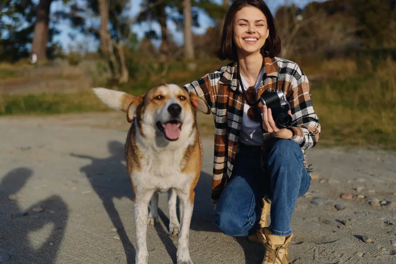 Woman with camera kneels beside a happy dog in outdoor nature, enjoying a casual walk on a sunny day, capturing candid moments of friendship, joy, and companionship during exploration.