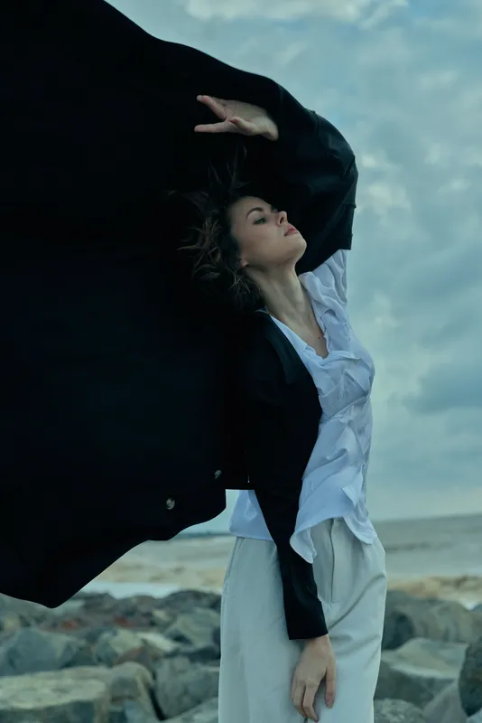 Woman in flowing white dress and black cape stands on a rocky beach, wind sweeping fabric for a dramatic fashion moment against sea and sky at dusk light
