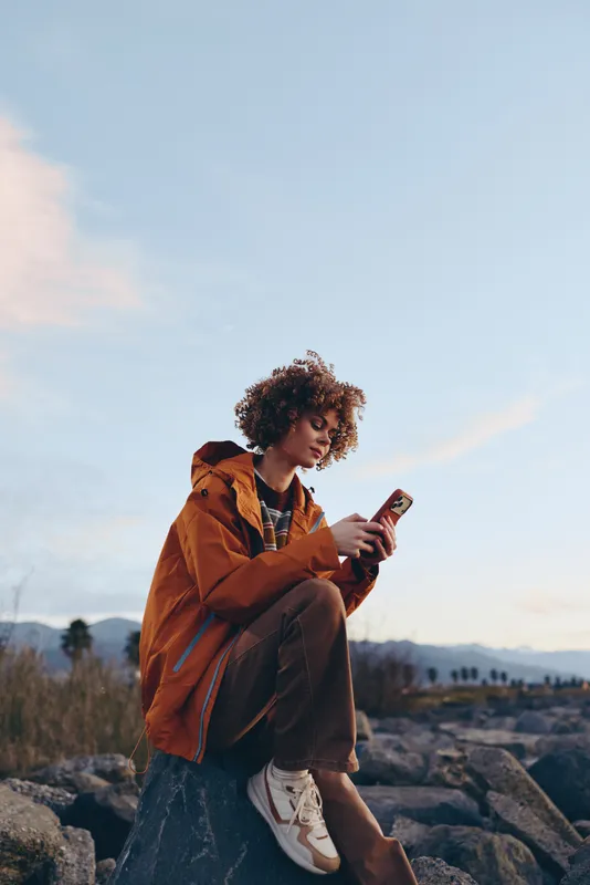 Woman lifestyle outdoor hiking portrait in orange jacket holding smartphone on rocky trail at sunset, distant mountains, travel mood, warm light, sky clouds