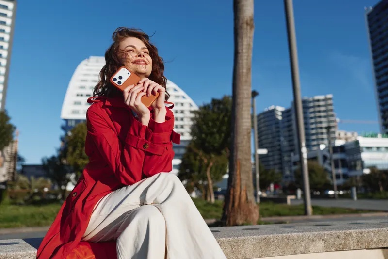 Young woman in a red coat sits outdoors on a sunny day holding a smartphone and smiling with closed eyes in an urban environment.