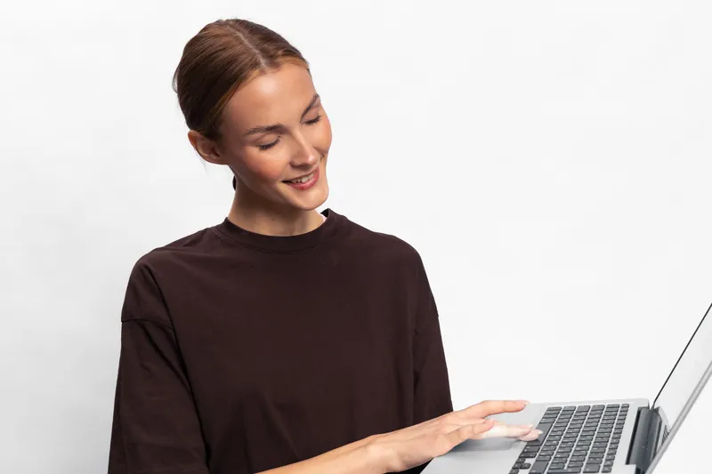 Young woman with tied hair wearing dark top smiles while holding laptop. Isolated on white background. Concept of technology, communication, and casual lifestyle with digital device.