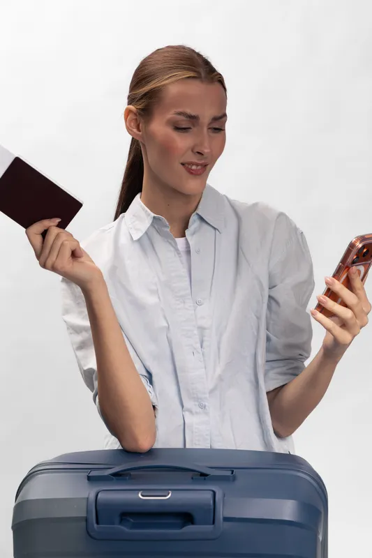 Confused young woman holds passport and smartphone near suitcase, wearing white shirt. Concept for travel uncertainty, planning, or ticket issues at airport or trip preparation.