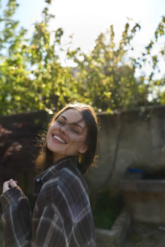 A smiling young woman in a plaid shirt stands outdoors in a sunlit yard, turning toward the camera with a warm expression