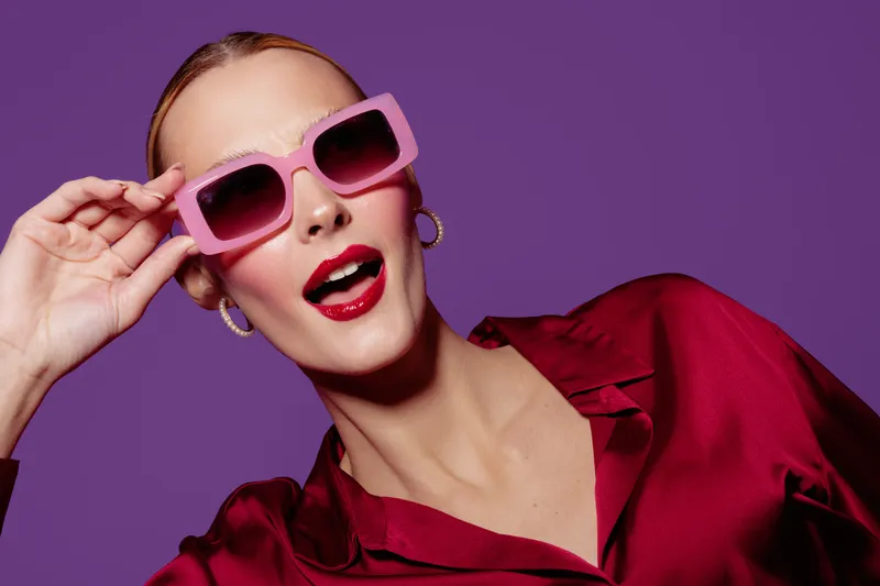 Stylish woman with pink sunglasses and red lipstick poses against purple background wearing red satin blouse. Concept of fashion, glamour, beauty, and confident attitude in studio.